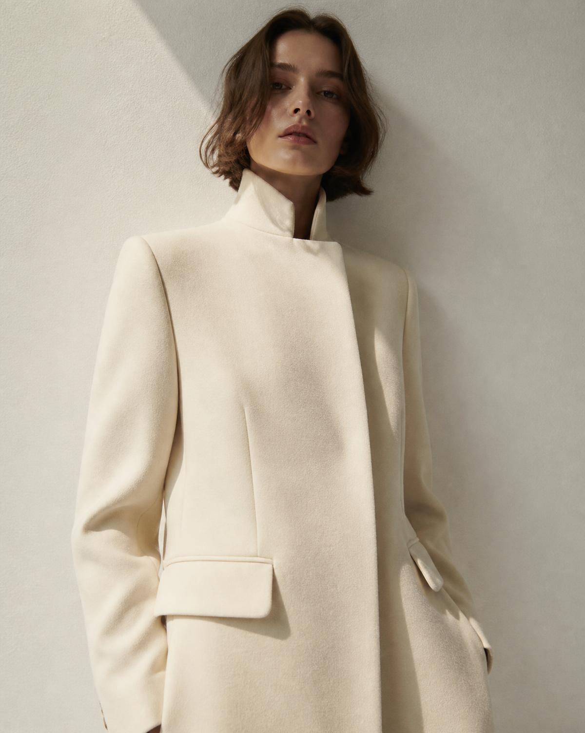 Model in a cream tailored coat against a white wall